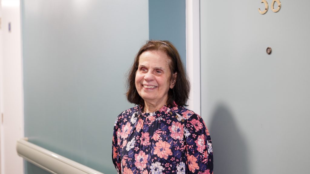 A woman stands in front of her apartment door, smiling. She's looking slightly to the left of camera.
