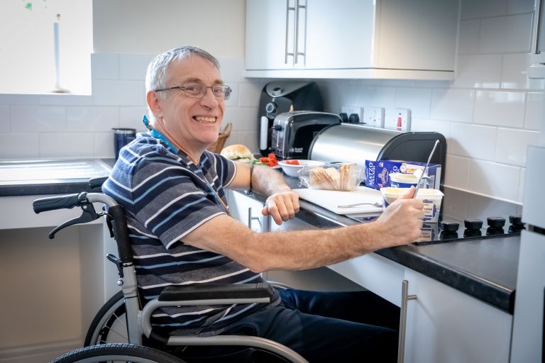 photo shows a tenant sat in his wheelchair at a kitchen worktop holding a knife in front of tub of butter