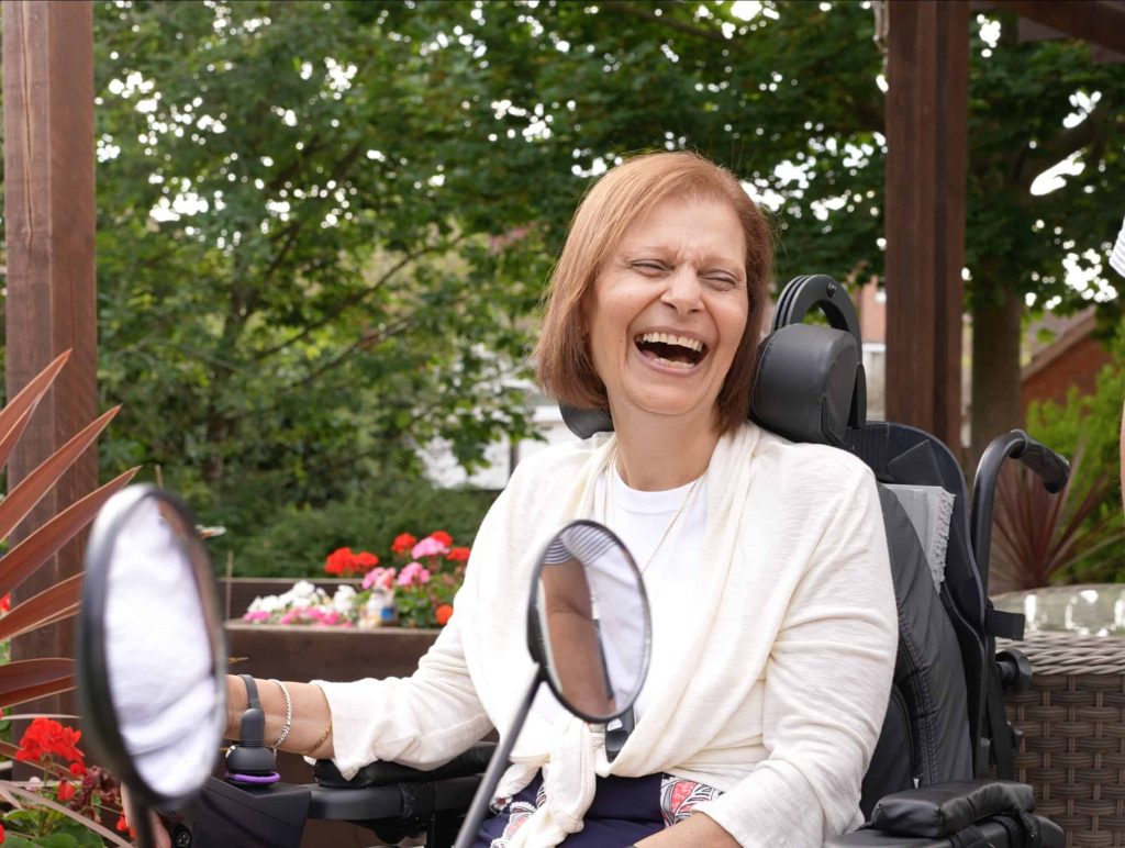 A woman laughs with a broad smile, and her eyes closed, she's sitting in a wheelchair in a garden