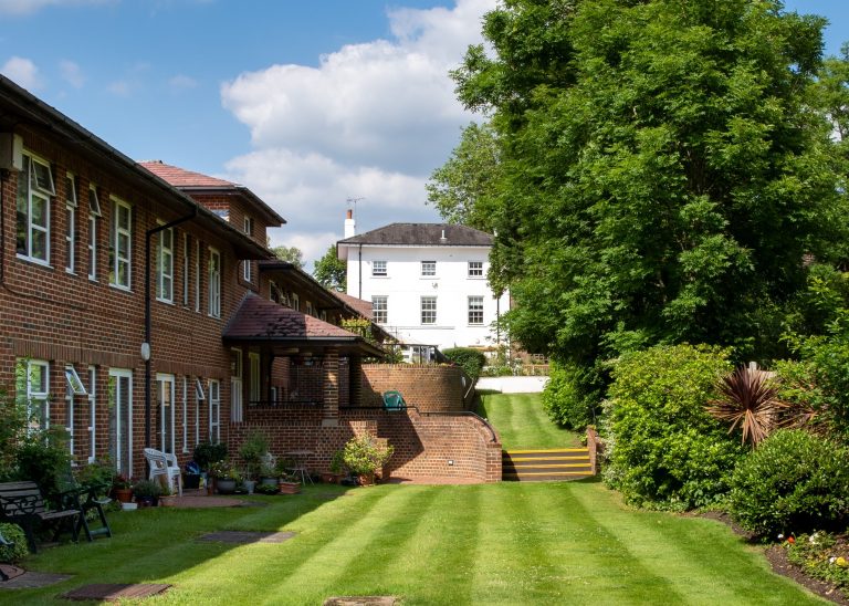 a white Georgian 3 storey house and a modern brick built 2 storey building stand in a garden with a green lawn