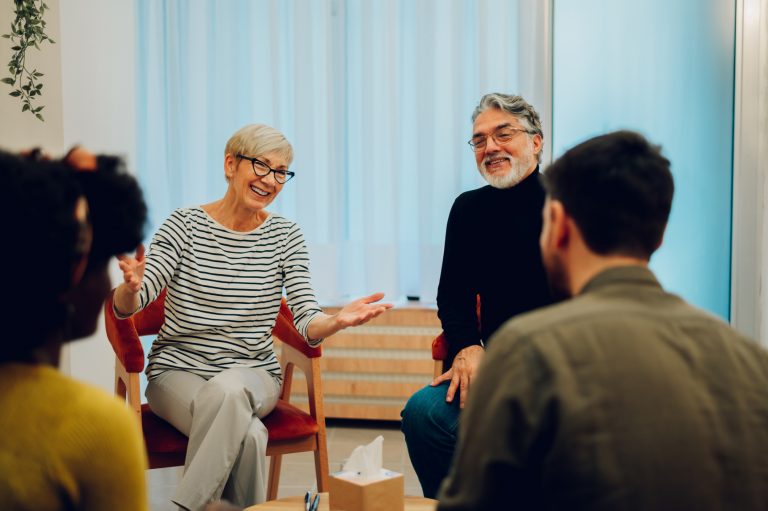 A group of 4 people sit on chairs in a circle A woman wearing glasses smiles, her arms are outstretched and palms face upwards.